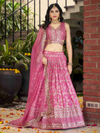 Woman in a pink traditional outfit standing outdoors with plants and a brick wall in the background.